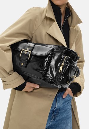 Persona con gabardina beige y vaqueros azules sosteniendo un bolso grande de cuero negro brillante con hebillas doradas y detalles de cremallera.