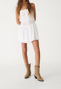 Vestido blanco sin mangas con textura fruncida y detalles de botones, combinado con botas de vaquero de ante marrón que presentan un bordado decorativo.