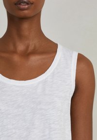 Bílý bezramínkový top s kulatým výstřihem, vyrobený z lehké, mírně strukturované tkaniny, která předvádí ležérní design.