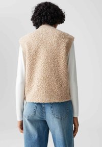 Gilet beige fluffy con colletto a coste, indossato sopra una camicia bianca a maniche lunghe. Abbinato a jeans in denim azzurro chiaro. Il finish testurizzato è ben evidente.