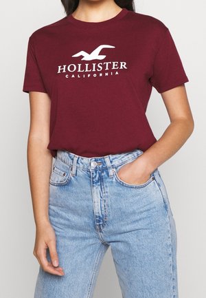 Burgunderfarvet bomulds T-shirt med rund hals og hvid "Hollister California" logo. Kombineret med lyseblå højtaljede jeans.