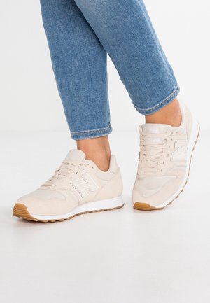 Persoon die lichtbeige New Balance-sneakers met rubberzolen en blauwe spijkerbroek draagt, staand op een witte vloer.