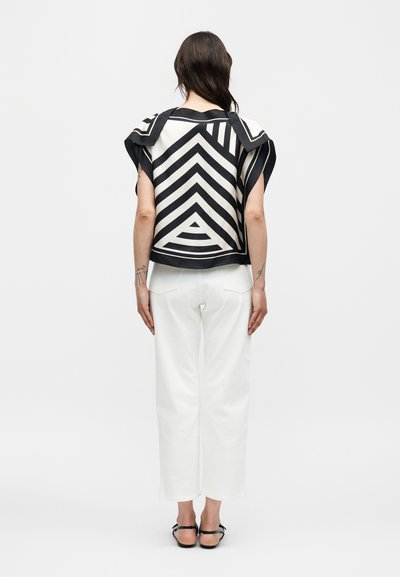 Γυναίκα με μαύρο και λευκό τοπ με γεωμετρικό μοτίβο και κοντά μανίκια, λευκό παντελόνι και μαύρα σανδάλια, στέκεται με την πλάτη γυρισμένη σε μονόχρωμο φόντο.
