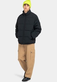 Giacca a pescatore nera con chiusura a zip, caratterizzata da un motivo a righe orizzontali e tasche laterali. Abbinata a pantaloni cargo color khaki e un berretto verde neon.