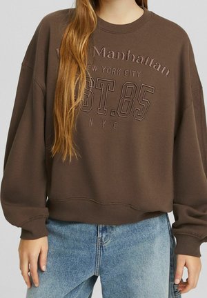 Sudadera marrón de gran tamaño con el texto "Manhattan New York City EST. 85 NYC", usada con vaqueros azul claro por una persona con el pelo largo.
