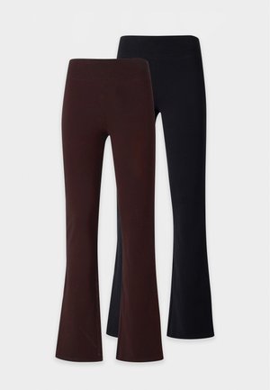 Frau mit geflochtenem Haar, die ein weißes Langarmshirt, dunkelbraune ausgestellte Hose und schwarze spitze Schuhe trägt, steht vor einem weißen Hintergrund.