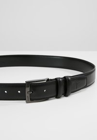 Cinturón de cuero negro con un acabado suave, que presenta una hebilla de metal y cuatro agujeros ajustables. Sutil logotipo embellecido en la correa.