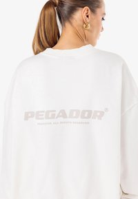 Fehér, nagyméretű pulóver, texturált anyagból készült. A hátsó részen egy nagy, világosszürke "PEGADOR" logó található, bordás mandzsettákkal és nyakkal.