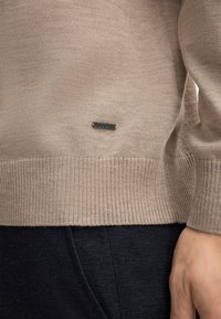 Beige Strickpullover mit geripptem Saum. Verfügt über ein kleines metallisches Logo-Tag. Wird über dunklen, strukturierten Hosen getragen, die glattes Material und Form zeigen.
