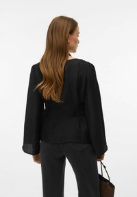 Melna teksturēta blūze ar vaļēju fitu, garām piedurknēm un savāktām detaļām. Kombinēta ar tumšām džinsa biksēm un brūnu somiņu.