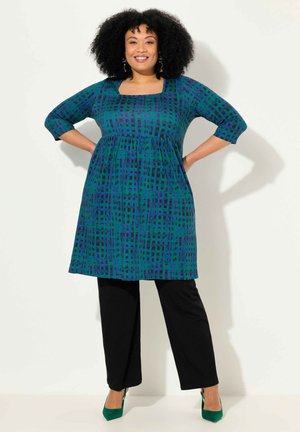 Grün- und blau gemustertes Kleid mit quadratischem Ausschnitt, Dreiviertelärmeln und einem gerafften Taillenbereich, kombiniert mit schwarzen Hosen und grünen Absätzen.