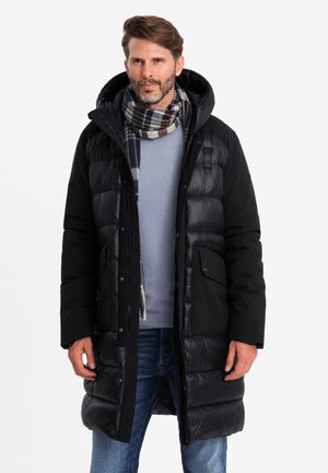 Manteau matelassé noir avec capuche, doté de poches zippées, mélange de panneaux matelassés et unis, porté sur un pull bleu et une écharpe à carreaux.