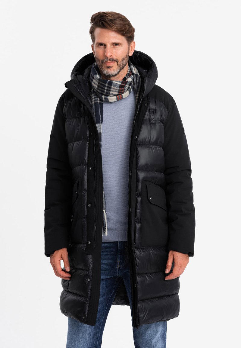 Schwarze Pufferjacke mit Kapuze, ausgestattet mit Reißverschlusstaschen, einem Mix aus gesteppten und soliden Paneelen, getragen über einem blauen Pullover und einem karierten Schal.