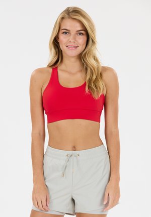 Frau mit langen blonden Haaren, die einen roten Sport-BH und hellgraue Shorts mit Kordelzug trägt, steht vor einem einfarbigen Hintergrund.