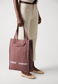 Bordeaux-farget stoff tote bag med to håndtak, med teksten "SAMSØE SAMSØE" i hvitt. Vesken har en rektangulær form og strukturelt design.