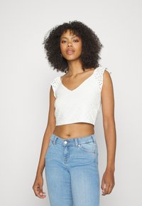 Top branco curto com detalhes de ilhós e alças com babados, combinado com jeans justos azul claro. O top possui um design de decote em V.