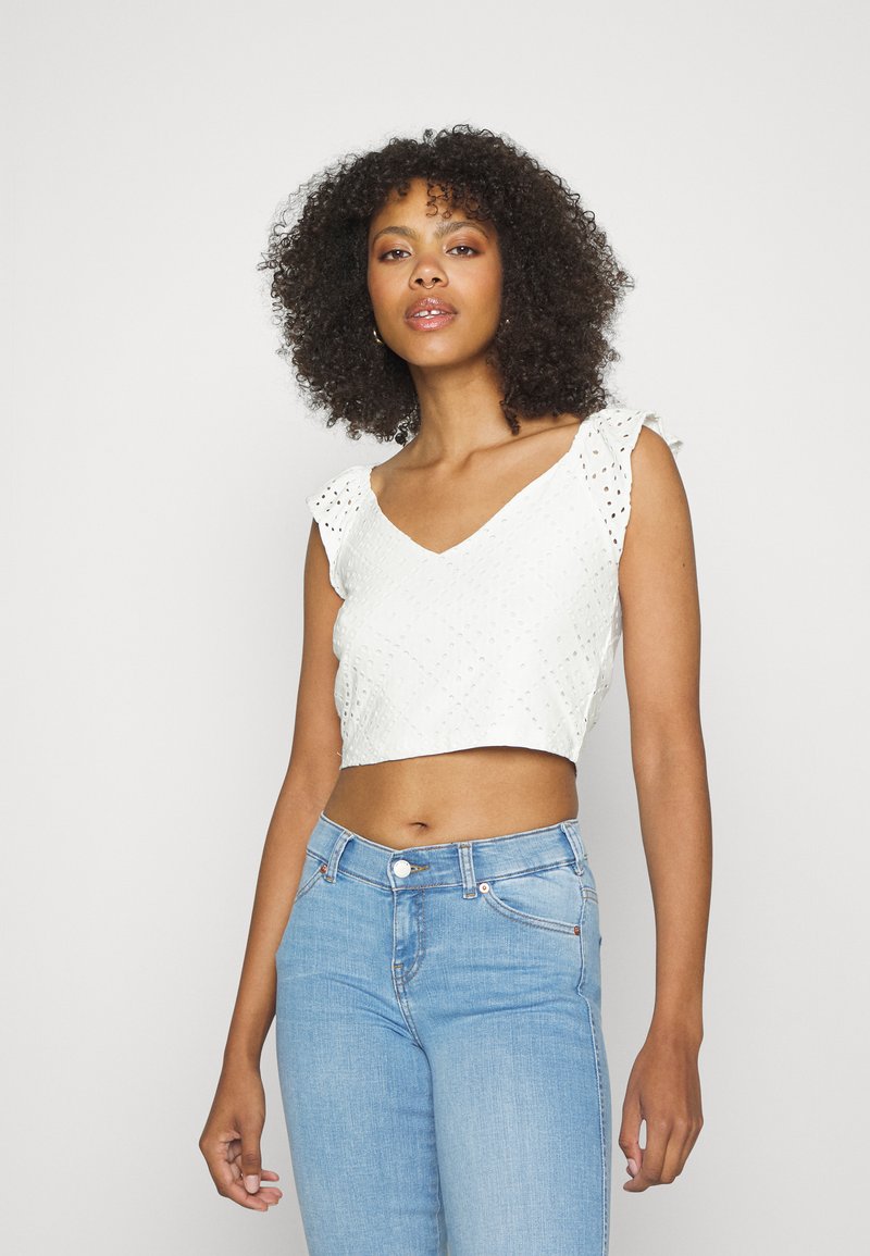 Top branco curto com detalhes de ilhós e alças com babados, combinado com jeans justos azul claro. O top possui um design de decote em V.