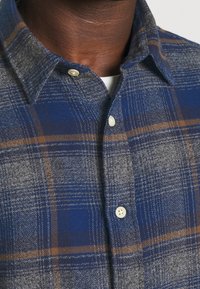 Blå och grå rutig skjorta med krage som knäpps, mjuk textur och ljusbruna accenter i mönstret. Två vita knappar synliga.