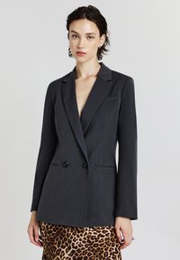 Femme jeune avec des cheveux courts et foncés, portant un blazer en charbon à double boutonnage et une jupe imprimée léopard, se tenant devant un fond blanc.