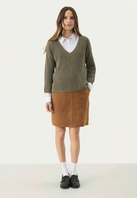 Oliwkowy sweter z prążkowanej dzianiny, w serek, nałożony na białą koszulę z kołnierzykiem, w połączeniu z brązową zamszową spódnicą do kolan i czarnymi loafersami.