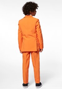 Orange jakkesæt med en tætsiddende design, der har en enkeltradet jakke og lige bukser, parret med sorte sneakers.