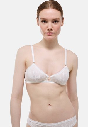 Femme à la peau claire et aux cheveux roux porte un soutien-gorge en dentelle blanche à motif floral et une culotte assortie, debout devant un fond clair uni.