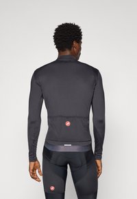 Mann in eng anliegendem schwarzem Langarm-Radtrikot und gepolsterten Radhosen, der mit dem Rücken zu einem schlichten Hintergrund steht.