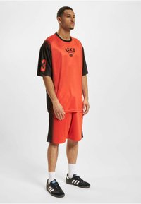 Rød og sort atletisk outfit bestående af en kortærmet skjorte med logo og nummer 3, parret med matchende shorts. Materialet virker let.