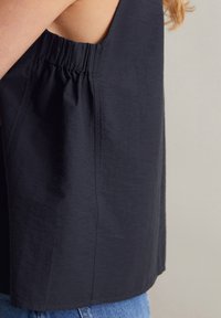 Mouwloos topje in marineblauwe stof met een geplooid naaddetail bij de halslijn, en een rechte snit die tot heupniveau reikt.