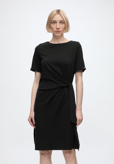 Femme aux cheveux blonds courts portant une robe noire à manches courtes avec un détail noué à la taille, debout devant un fond clair uni.