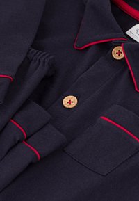 Navy katoenen poloshirt met rode bies langs de kraag en zak. Kenmerkt zich door houten knopen en een borstzak voor extra detail.