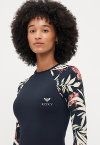 Roxy long-sleeve swimsuit featuring a navy body, floral-patterned sleeves in shades of pink and green, and a small logo on the chest.