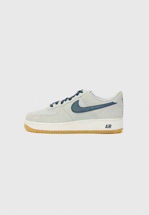 Jasnoszare buty Nike Air Force 1 z ciemnoniebieskim logo i sznurowadłami, białą środkową częścią podeszwy i gumową podeszwą na jednolitym tle.
