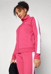 Roza zip-up trenirka ima bele stranske črte, visok ovratnik in prilegajoče manšete. Gladka tekstura tkanine z atletskim dizajnom.