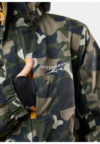 Hand som drar en gul dragkedja på en grön och svart kamouflagejacka med Swedemount-logotyp och bergdesign på bröstet.