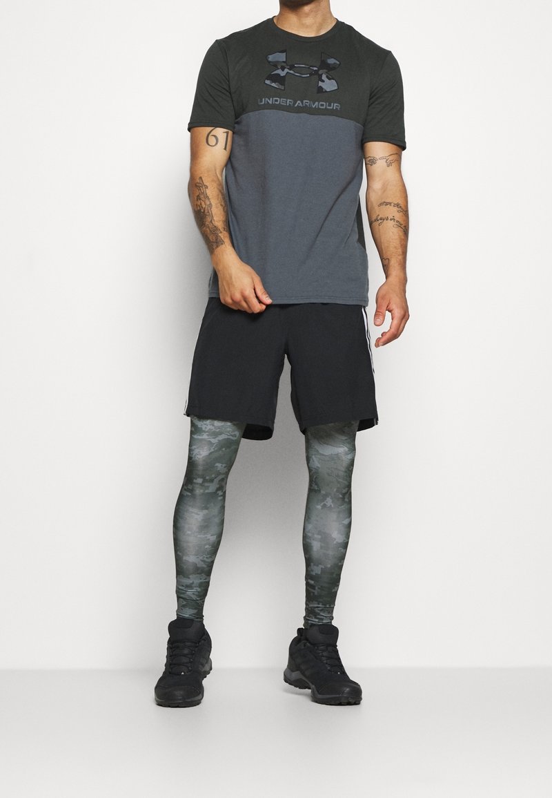 Pantalones cortos negros, camiseta gris con estampado de camuflaje y el logo de "Under Armour", mallas de camuflaje verde y zapatillas deportivas negras.