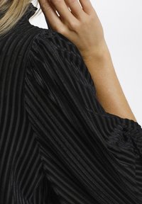 Svart randig blus med textil med lyster, puffärmar och hög krage. Ränderna är vertikala och smala.