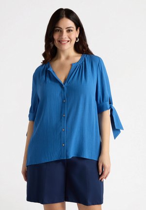 Blauwe blouse met knoopsluiting en een gestructureerde afwerking, met een V-hals, korte mouwen met strikjes en een relaxed fit. Draagt met donkere shorts.