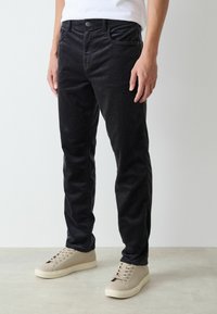 Black cotton trousers with a slim fit, featuring front pockets, button closure, and slightly faded texture. Paired with beige sneakers.