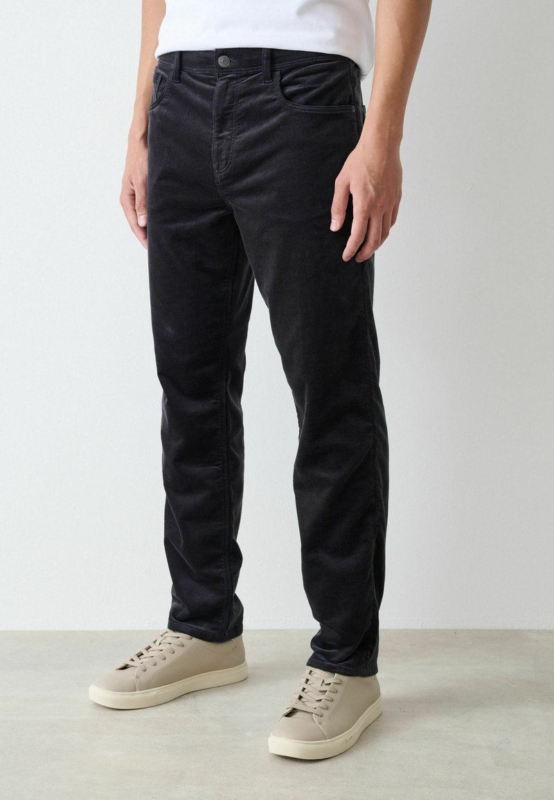 Black cotton trousers with a slim fit, featuring front pockets, button closure, and slightly faded texture. Paired with beige sneakers.