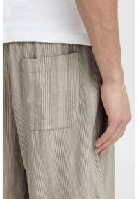 Gestreifte beige Hose aus strukturiertem Stoff, mit einer Gesäßtasche und lockerer Passform. Sichtbare Hand ruht an der Seitennaht.