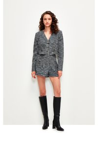 Blazer de tweed gris con botones dorados, combinado con pantalones cortos a juego que cuentan con bolsillos y detalles dorados. Botas negras hasta la rodilla completan el look.