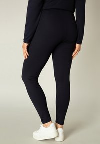 Marineblauwe leggings van rekbare stof, aansluitend ontwerp en een gladde textuur. Gedirigeerd vanuit een achteraanzicht, gedragen met witte platform sneakers.