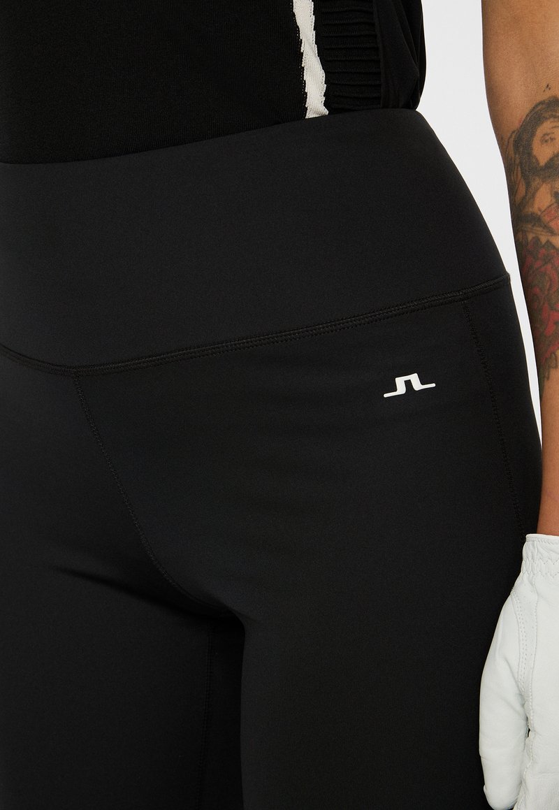 Närbild av svarta högmidjade leggings med vitt logotyp nära höften, som bärs med en svart topp och en vit handske på en tatuerad arm.