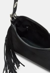 Sort suedette clutchtaske med lynlås, indvendig lomme og dekorativt frynse-detalje. Indersiden er foret med et blødt sort stof.