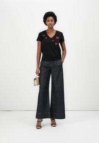 Schwarzes V-Ausschnitt-T-Shirt mit roter Herzstickerei, kombiniert mit weiten dunkelblauen Jeans, ergänzt durch schwarze Sandalen mit Riemchen und einer kleinen beigen Tasche.