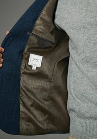Blaues Fischgrätmuster-Jacket mit einem glatten braunen Futter mit subtilen Mustern, Innentasche und einem Etikett mit der Aufschrift "NEXT Tailoring."