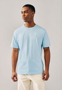 Hellblaues Baumwoll-T-Shirt mit klassischem Rundhalsausschnitt, kurzen Ärmeln und kleinem besticktem Logo auf der Brust. Schlichte, glatte Textur.