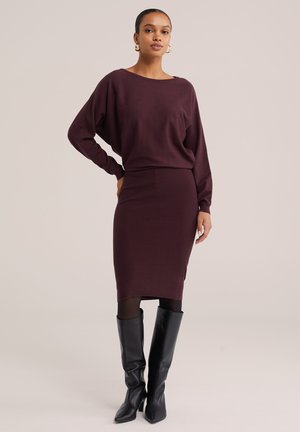 Femme portant un haut à manches longues violet foncé et une jupe assortie jusqu'au genou, des collants noirs et des bottes noires montant jusqu'au genou, debout avec une main sur la hanche.