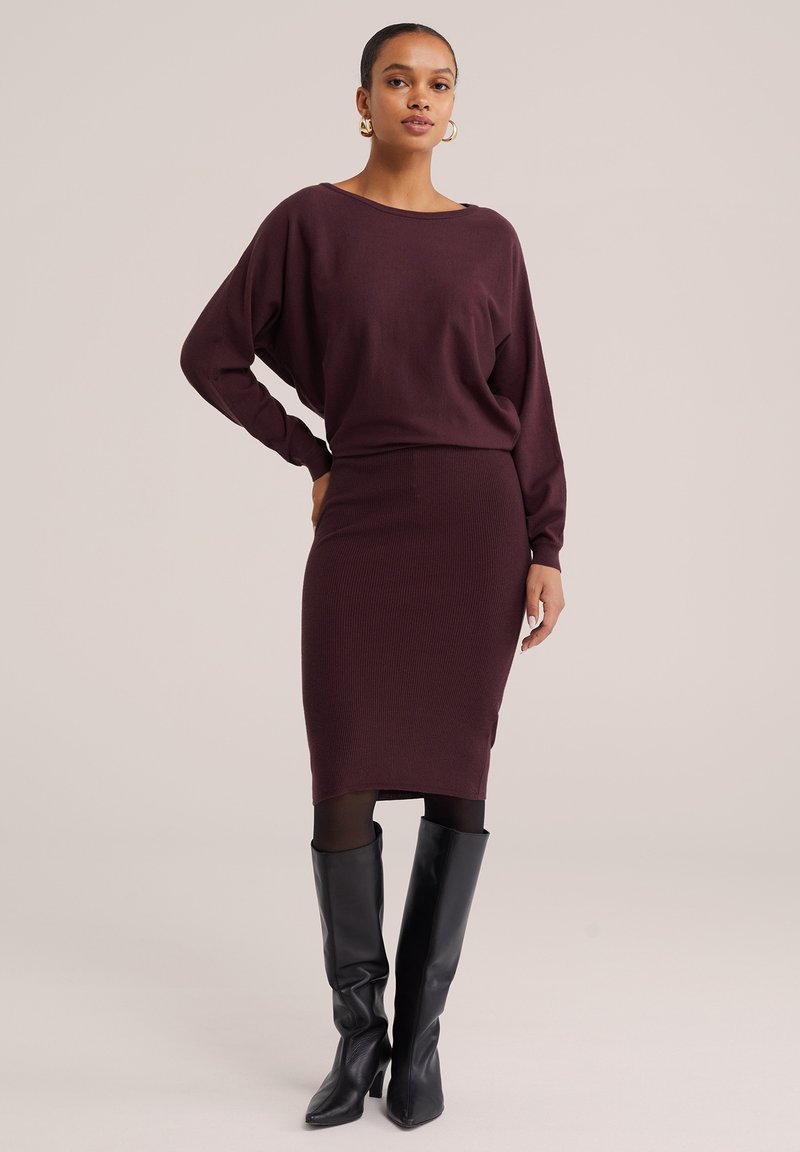 Femme portant un haut à manches longues violet foncé et une jupe assortie jusqu'au genou, des collants noirs et des bottes noires montant jusqu'au genou, debout avec une main sur la hanche.